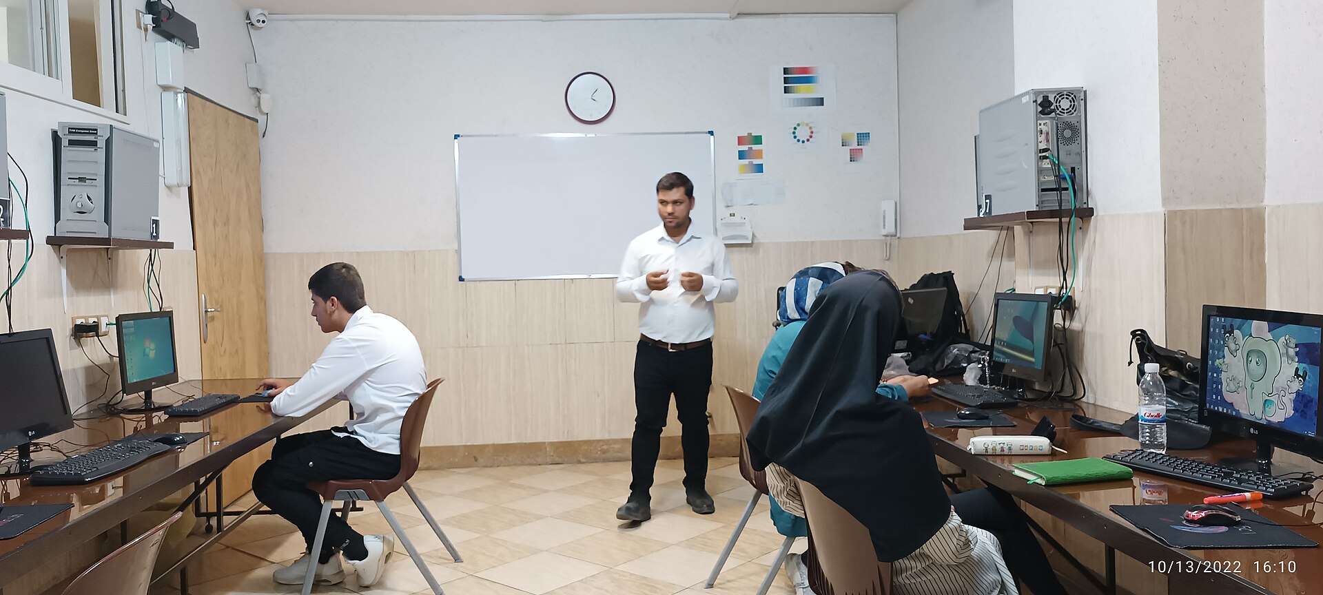 Professor orientando aluno diante de um computador em ambiente educacional — foto de Masoud mahfouzi, Wikimedia Commons, CC BY-SA 4.0.