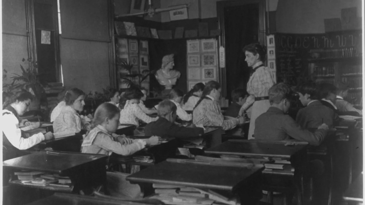 Professora acompanha uma turma durante uma atividade em sala. Fotografia histórica de uso público, via Wikimedia Commons.