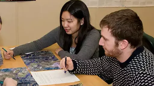 Dois estudantes colaboram à mesa, discutindo e registrando uma atividade de aprendizagem.